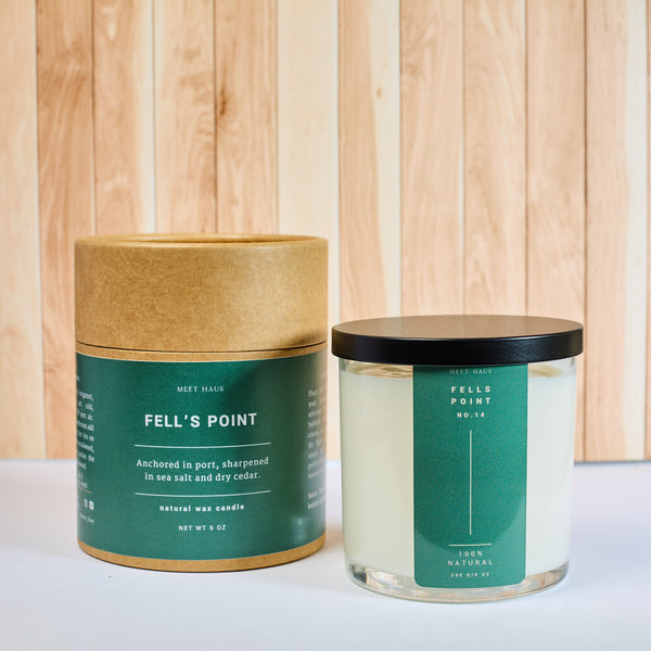 Meet Haus Fells Point soy candle, a Baltimore neighborhood scent: 9 oz clear glass jar with black lid and green label beside its kraft tube box noting sea salt and dry cedar, on white against pale wood panels.