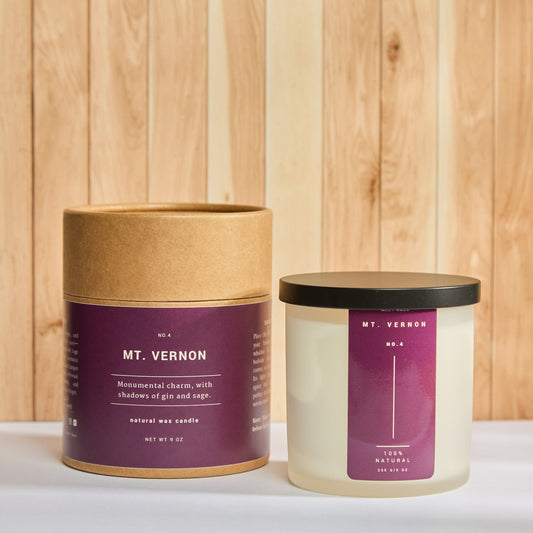 Frosted glass candle labeled “MT. VERNON” beside its kraft tube packaging; deep plum label reads: “Monumental charm, with shadows of gin and sage.