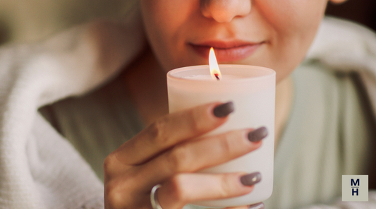 Close-up of a woman holding a lit white candle near her face, smiling softly as she enjoys the fragrance.