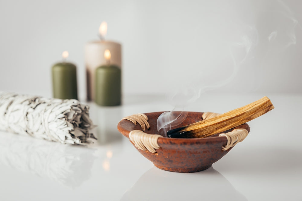A smoking stick of palo santo rests in a rustic clay bowl on a white surface, with lit sage, green and beige candles softly glowing in the background.