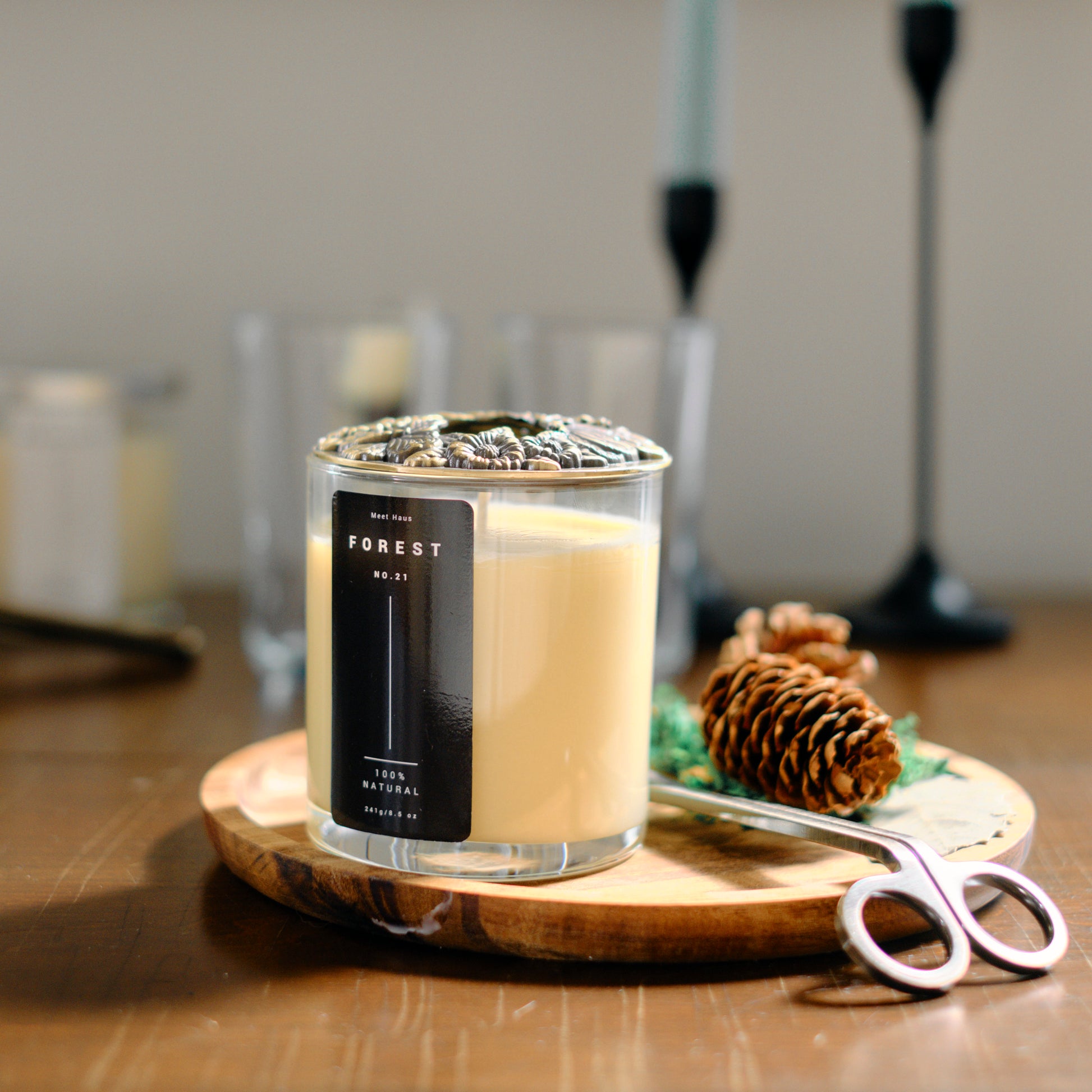 A Meet Haus Forest No. 21 candle, with a pine cone and a pair of scissors on a wooden tray, is set against a blurred background featuring elegant candle holders.