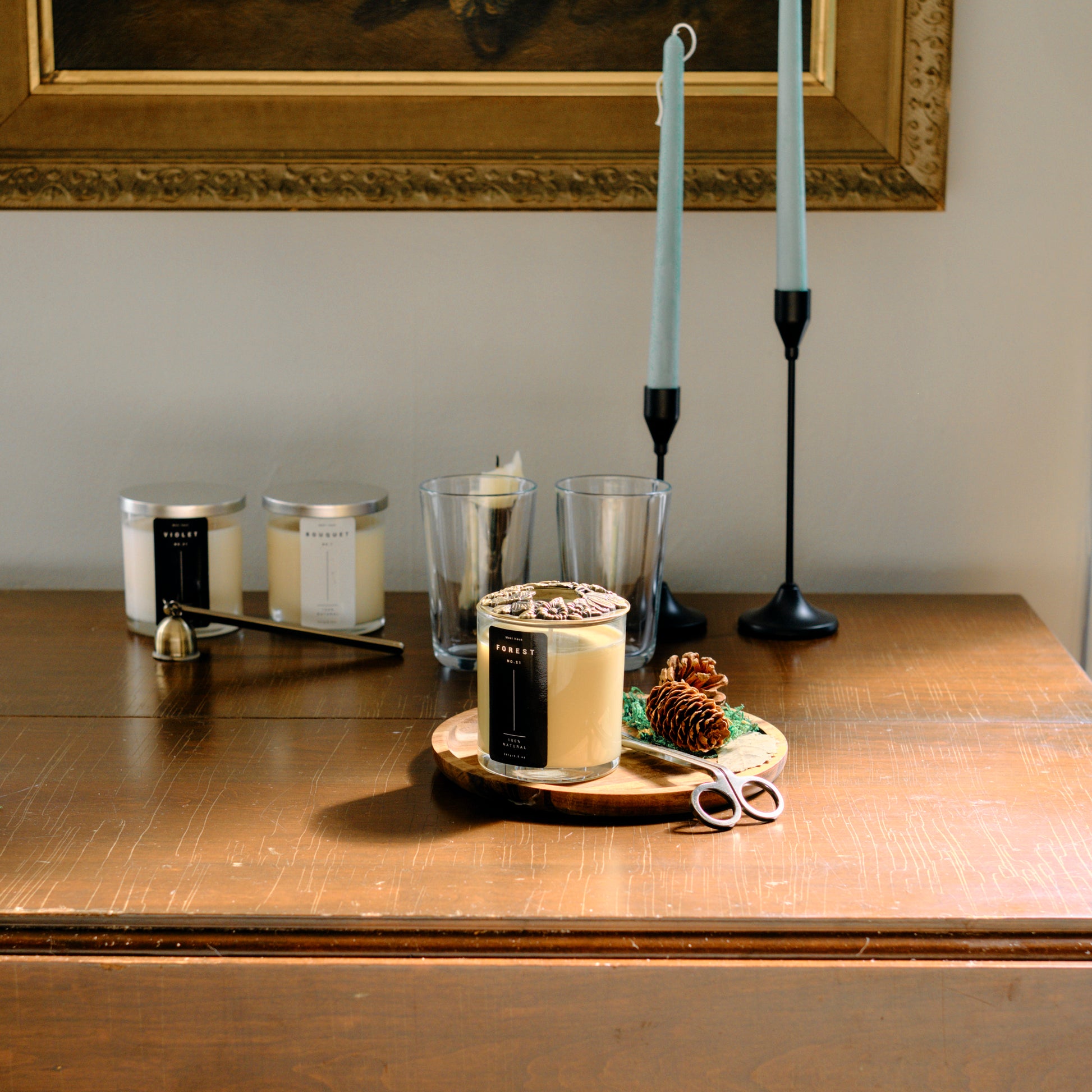 An array of Meet Haus candles on an antique table, featuring the prominent Forest candle with pine cones and scissors, flanked by elegant tall candlesticks and a decorative frame in the background.