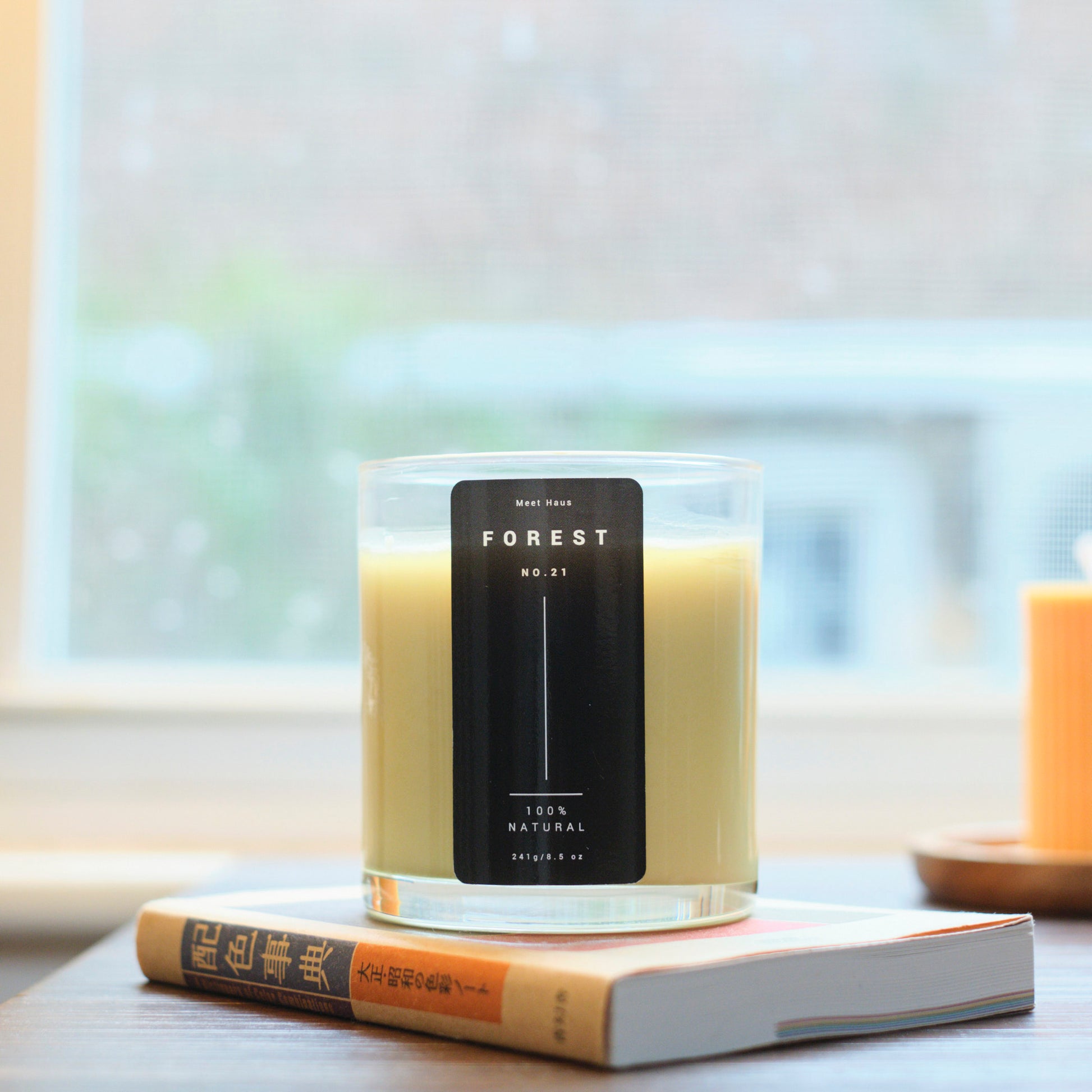 A lit Meet Haus Forest No. 21 soy candle sits atop a book on a wooden table by a window with a view of greenery outside, inviting a cozy reading atmosphere.