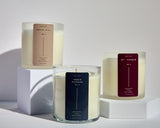 Three Meet Haus candles displayed on white blocks, featuring the Druid Hill candle with a tan label, the Inner Harbor candle with a navy label, and the Mt. Vernon candle with a burgundy label. Each candle is in a clear glass jar with creamy white soy wax.