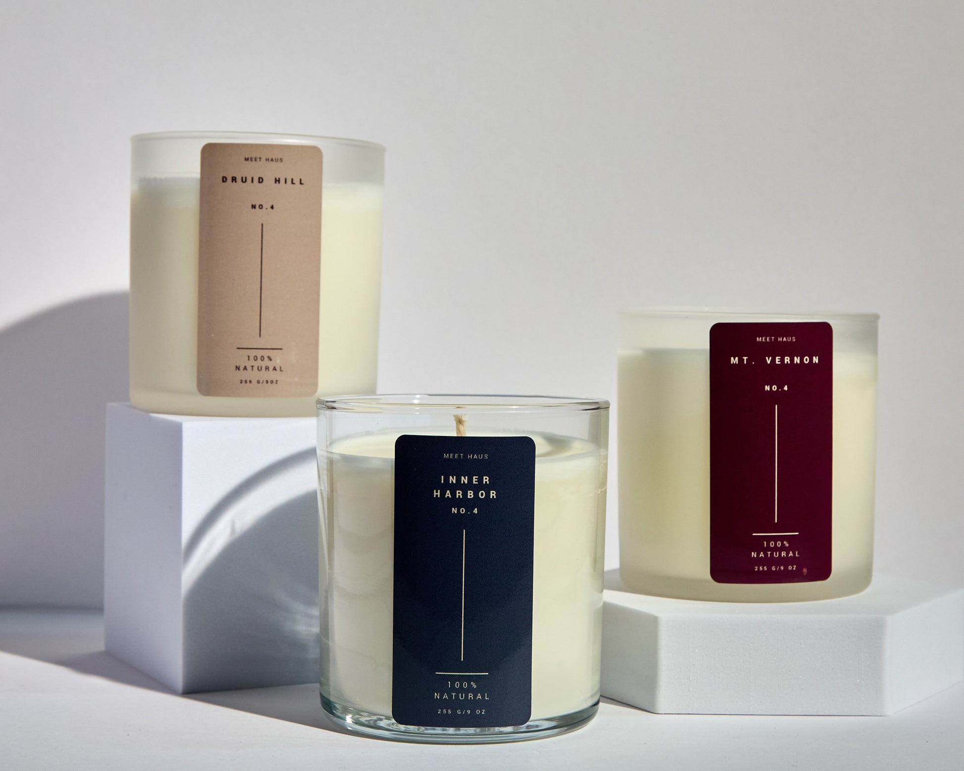 hree Meet Haus candles displayed on white blocks, featuring the Druid Hill candle with a tan label, the Inner Harbor candle with a navy label, and the Mt. Vernon candle with a burgundy label. Each candle is in a clear glass jar with creamy white soy wax.