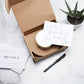 An open Meet Haus branded cardboard box on a marble countertop displays a neatly packaged candle, accompanied by candle care instructions and a 'Thank You' card from Meet Haus, with a sleek black pen and a potted succulent plant beside it.