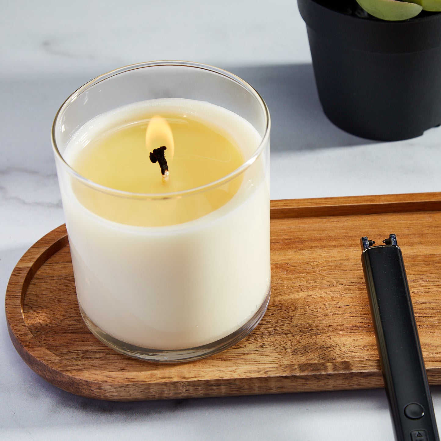 A lit Meet Haus Bouquet candle with a gently flickering flame, placed on a wooden tray next to a modern black lighter, with a succulent plant in the backdrop, set on a marble surface.