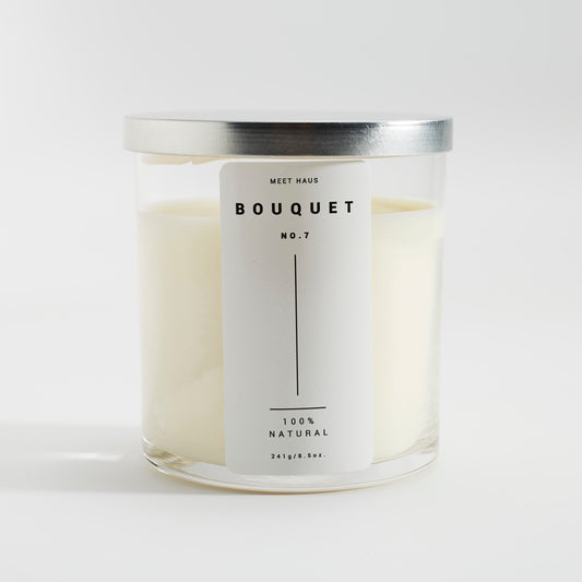 A Meet Haus Bouquet No.7 candle placed on a textured gray surface, featuring a minimalist label, with the candle wax visible through the clear glass container.
