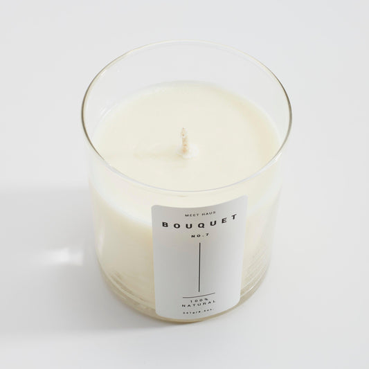 A Meet Haus Bouquet No.7 candle placed on a textured gray surface, featuring a minimalist label, with the candle wax visible through the clear glass container.