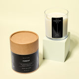 A clear glass Forest candle with creamy white wax sits on a pale green block beside its round kraft paper packaging tube. Both labels feature minimalist black design against a soft yellow background.
