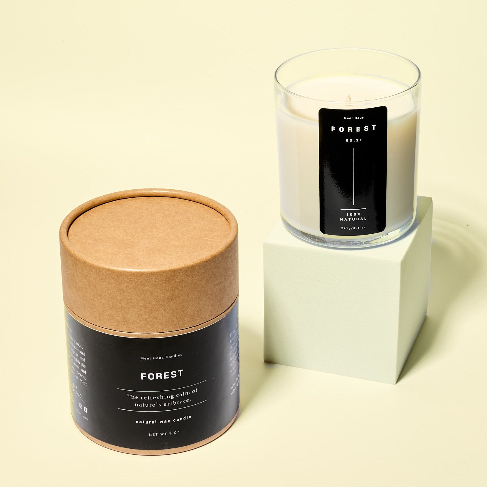 A clear glass Forest candle with creamy white wax sits on a pale green block beside its round kraft paper packaging tube. Both labels feature minimalist black design against a soft yellow background.