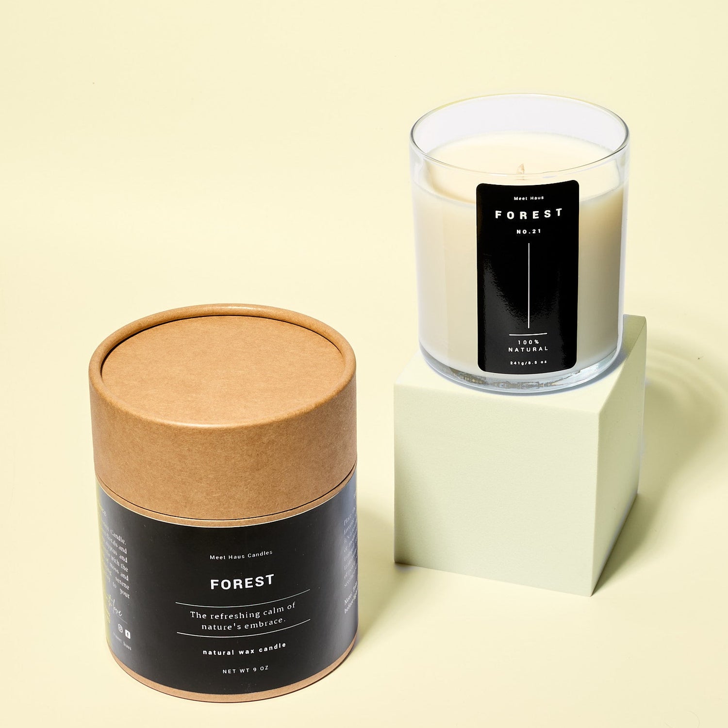 A clear glass Forest candle with creamy white wax sits on a pale green block beside its round kraft paper packaging tube. Both labels feature minimalist black design against a soft yellow background.